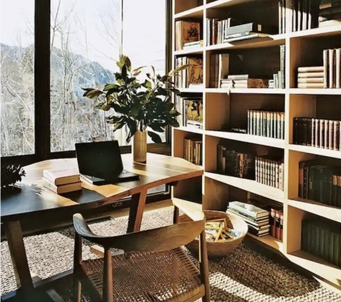 A sunlit reading room with bookshelves, a wooden desk, a laptop, and a woven chair.
